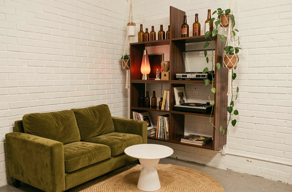 Petit salon urbain avec étagère en noyer, canapé en velours côtelé vert olive et accessoires vintage années 70 comme une lampe à lave et un tourne-disque